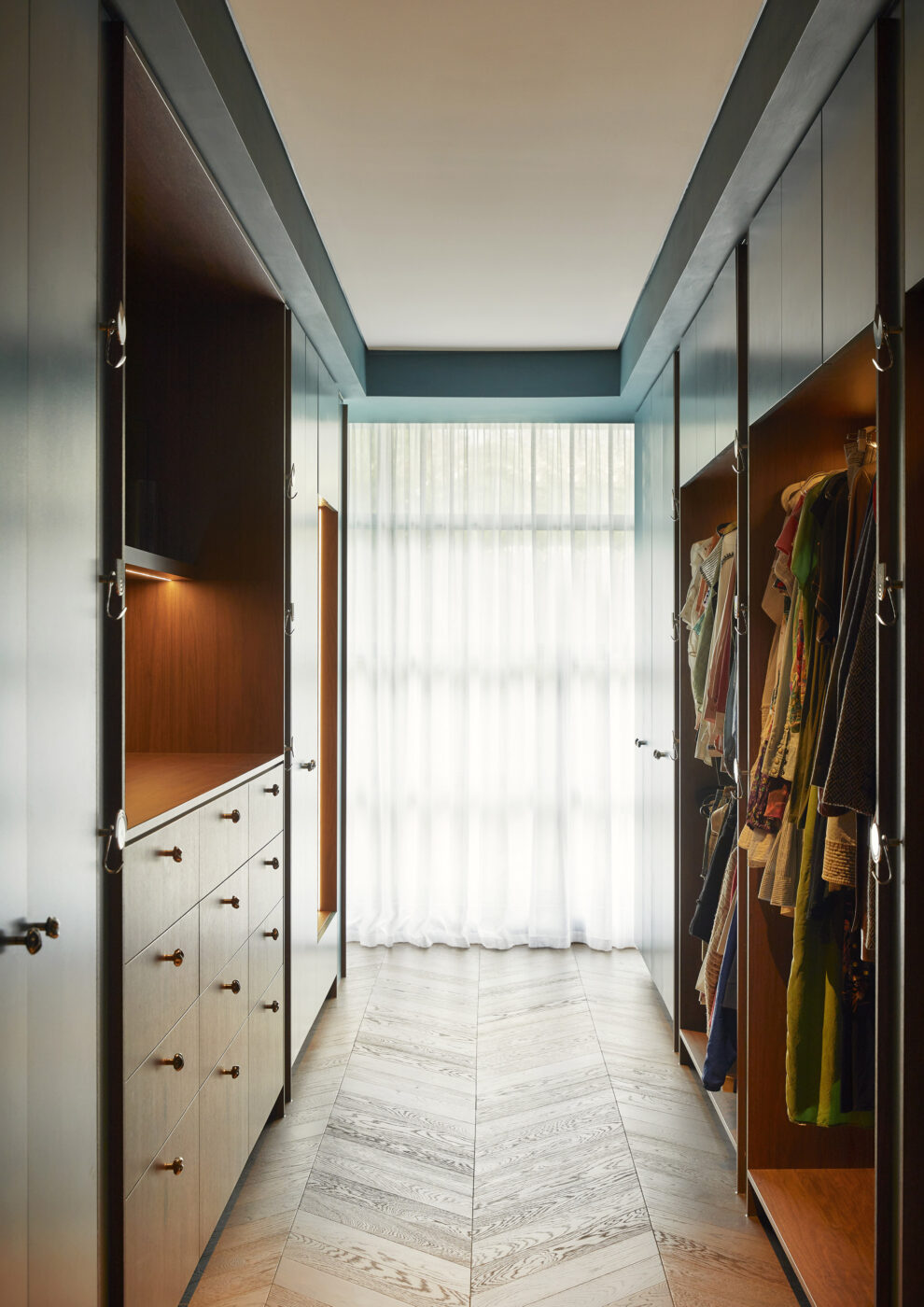 Designer walk-in wardrobe with bespoke teal-painted cabinetry, walnut veneer detailing and floor-to-ceiling linen sheer curtains, combining luxurious materials, texture and contemporary craftsmanship.