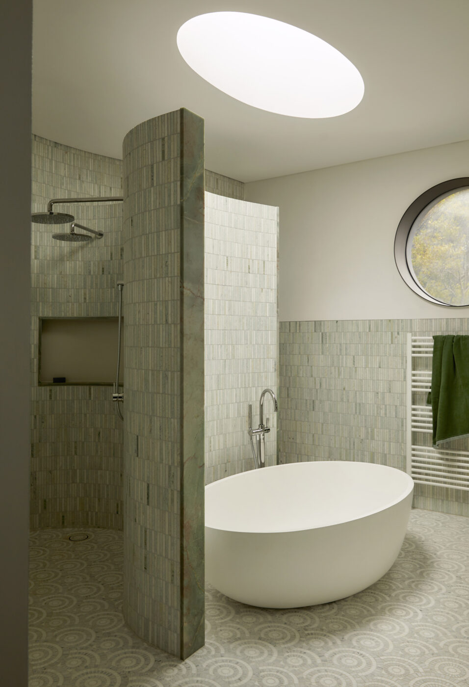 Architecturally designed circular ensuite with a freestanding bath under a round skylight, curved shower wall, celadon green marble wall tiles and patterned marble mosaic flooring, blending timeless materials with contemporary luxury.