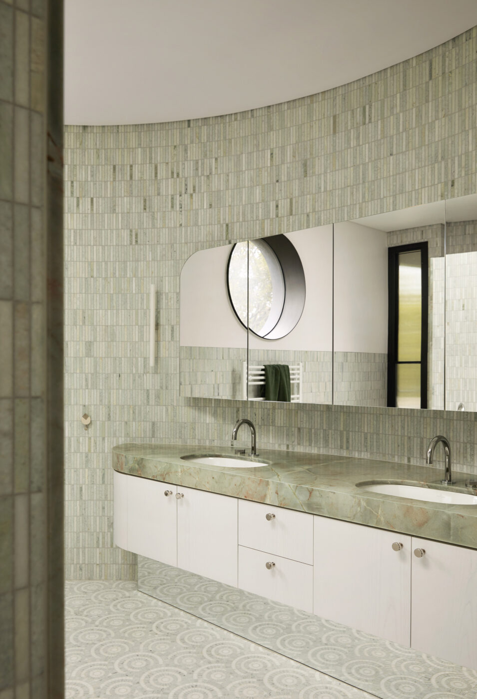 Designer ensuite in a curved architectural space with celadon green marble wall tiles, geometric grey and white floor tiles and a custom green stone vanity, blending natural materials with contemporary elegance.