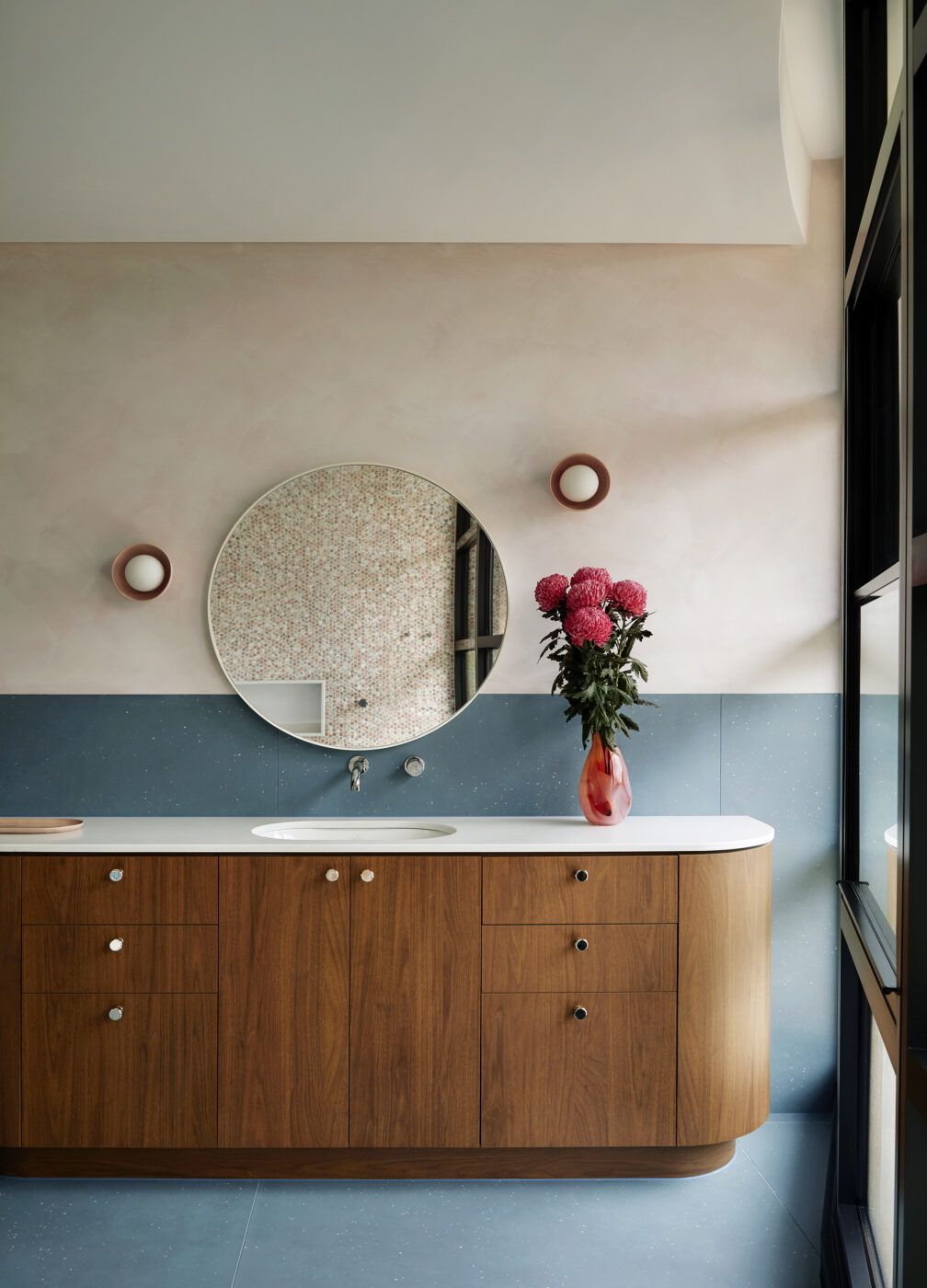 Designer ensuite bathroom with blush pink walls, statement blue tiles, custom curved timber veneer vanity, large circular mirror and decorative porcelain wall sconces, blending natural materials with contemporary elegance.