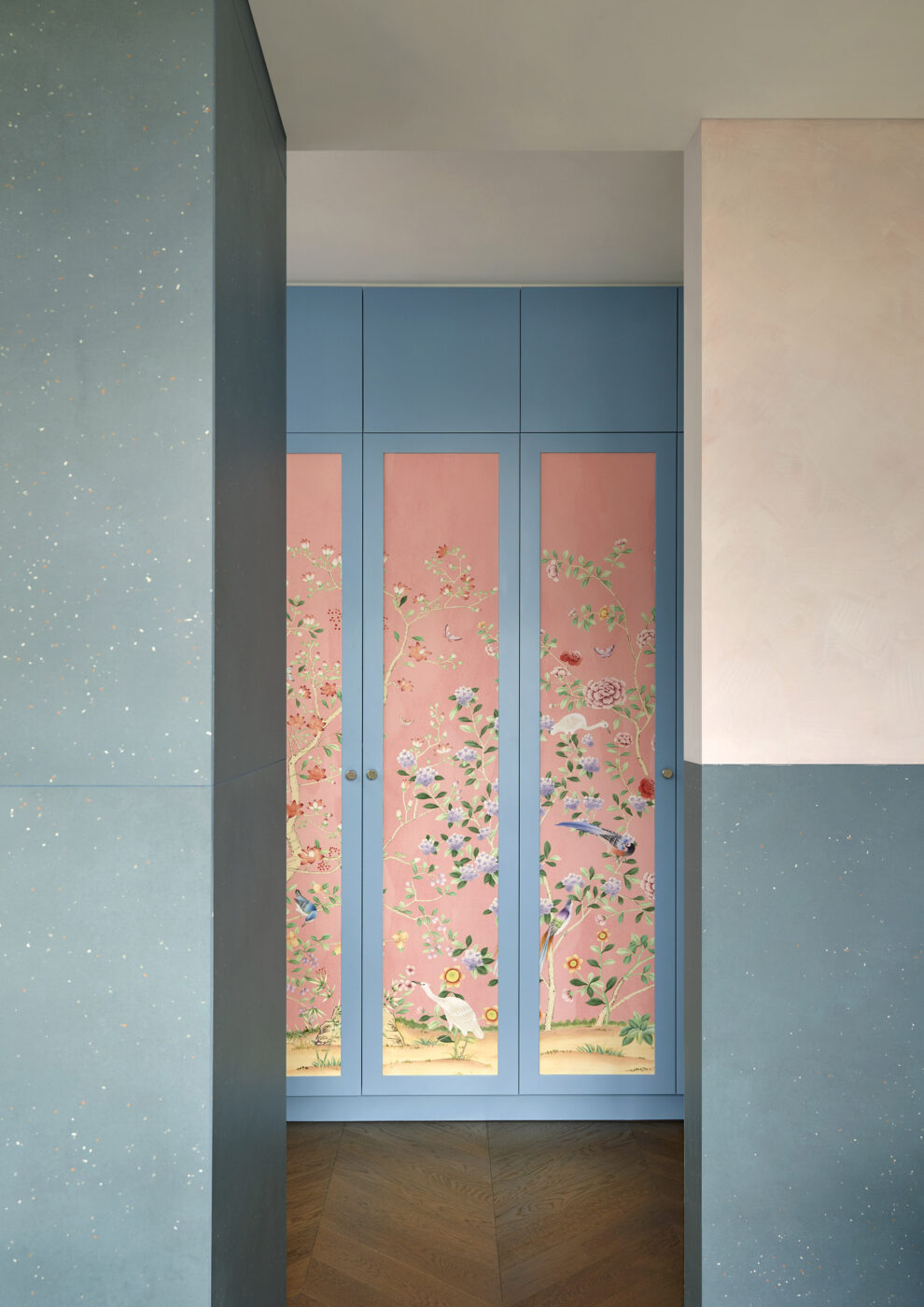 Luxury ensuite bathroom opening onto a custom walk-in wardrobe with blue painted doors and pink chinoiserie wallpaper detailing, blending bold colour, decorative pattern and contemporary interior design.