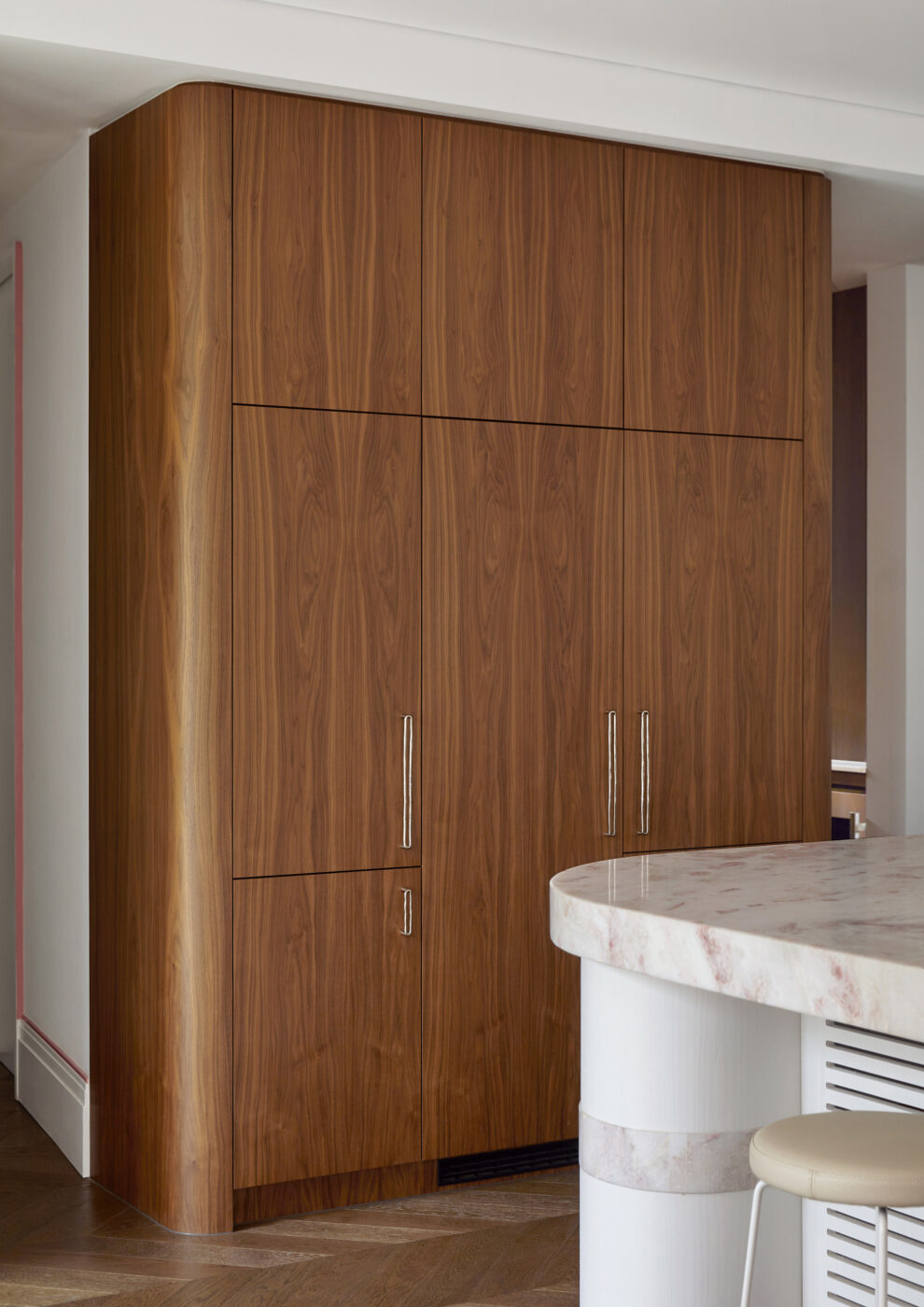 Luxury kitchen with tall walnut veneer cabinets housing an integrated refrigerator, bespoke solid brass handles in polished nickel and a statement island featuring a white veneer base and pink and white marble countertop.