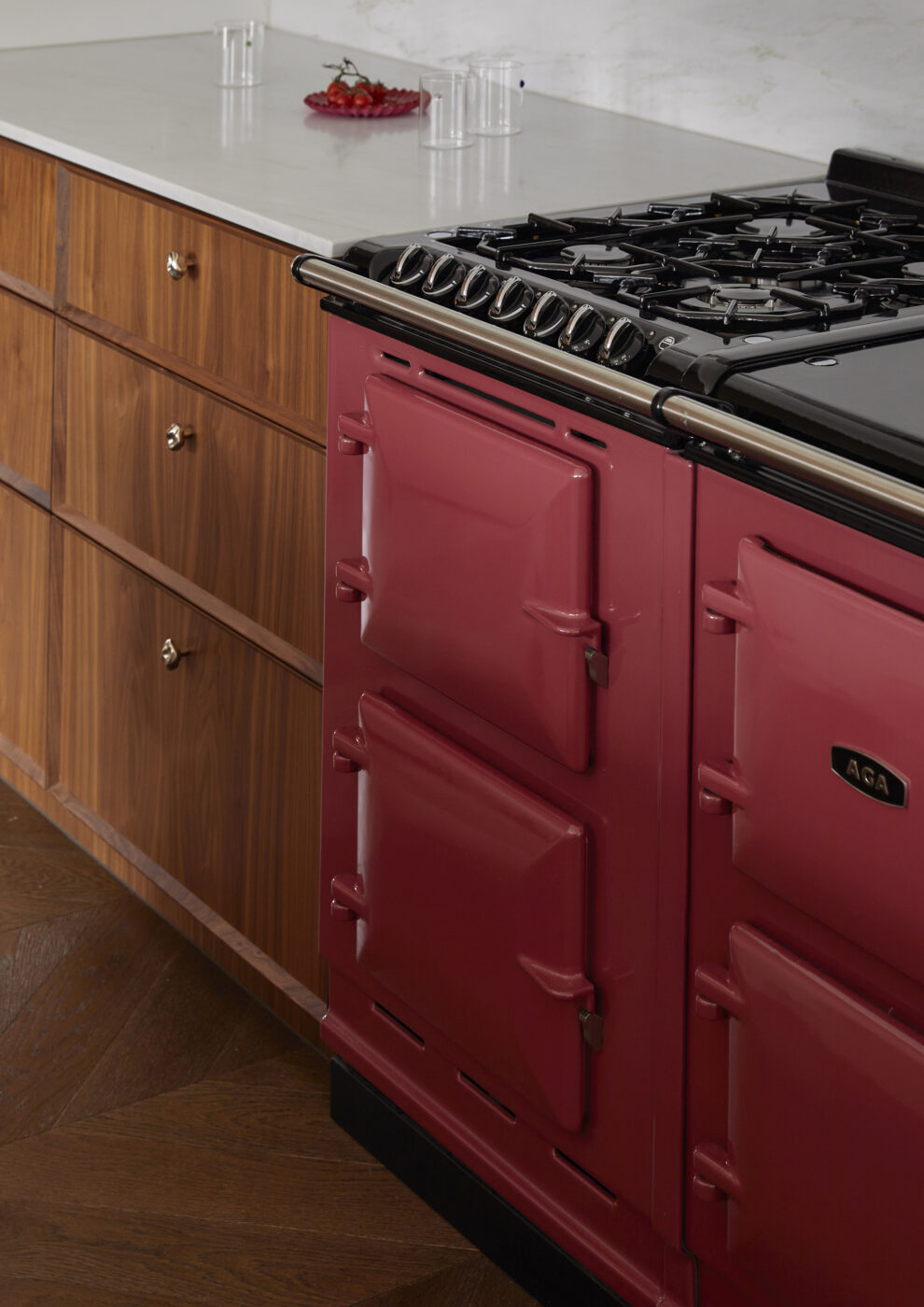 Designer kitchen detail highlighting a statement raspberry AGA cooker, custom walnut veneer cabinetry and bevelled drawer fronts, blending colour, texture and premium craftsmanship in a luxury kitchen.