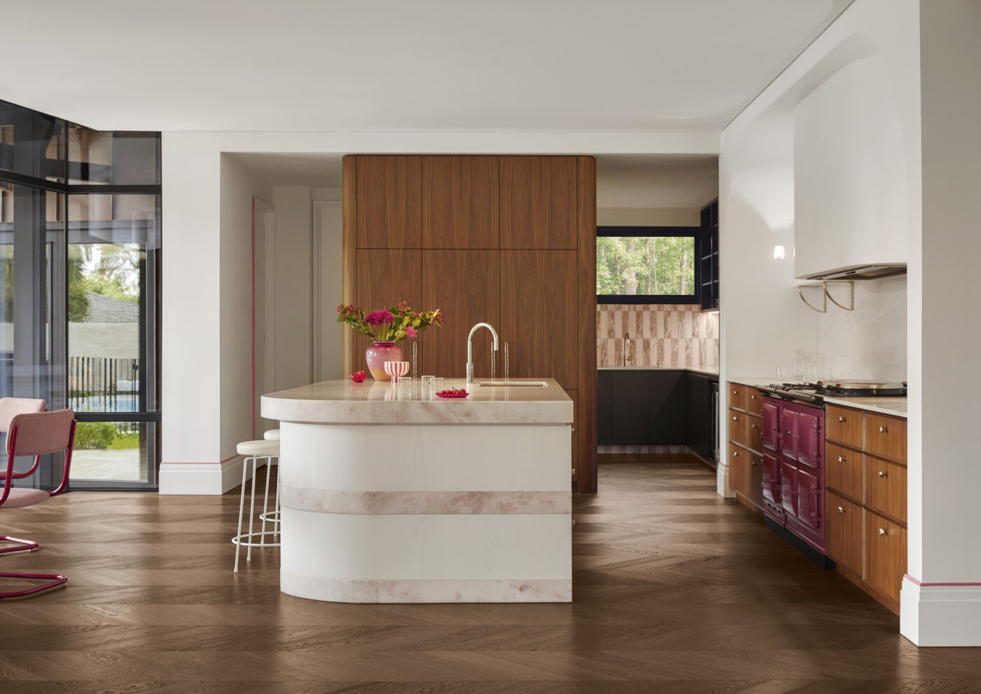 Luxury kitchen featuring a statement raspberry-coloured AGA oven, white marble island with soft pink veining and marble detailing wrapping the sides of the white veneer joinery. Walnut veneer tall cabinetry conceals integrated appliances, while a dark stained chevron timber floor adds warmth and contrast. Views through to the butler's pantry reveal pink and white marble checkerboard tiles, creating a sophisticated yet playful interior.